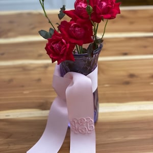 K&ouml;nnte beinhalten: Eine violette Glasvase mit mehreren dunkelroten Rosen und gr&uuml;nen Stielen. Ein hellrosa Band ist um den Hals der Vase gebunden und zeigt ein kreisf&ouml;rmiges, gesticktes Monogramm in passendem Rosaton auf dem herabh&auml;ngenden Ende des Bandes.