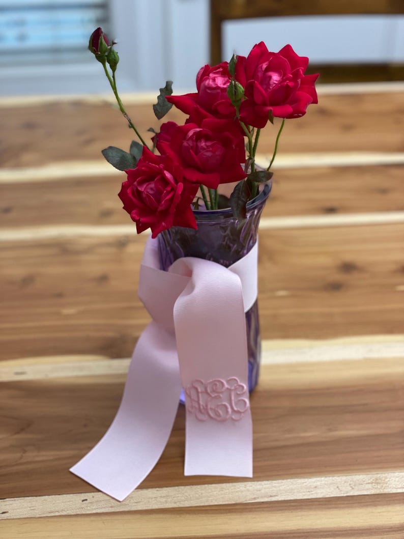 K&ouml;nnte beinhalten: Eine violette Glasvase mit mehreren dunkelroten Rosen und gr&uuml;nen Stielen. Ein hellrosa Band ist um den Hals der Vase gebunden und zeigt ein kreisf&ouml;rmiges, gesticktes Monogramm in passendem Rosaton auf dem herabh&auml;ngenden Ende des Bandes.