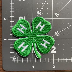 May include: A green four-leaf clover patch with a white embroidered border and four white letter H characters, one on each leaf. The patch is positioned on a dark gray cutting mat with white grid lines and markings, measuring approximately 2 inches wide.