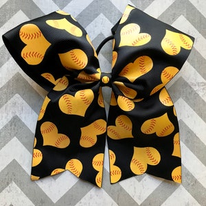 May include: Black and yellow softball-themed hair bow with a red stitching design. The bow is made of a soft, textured fabric and has a heart shape.