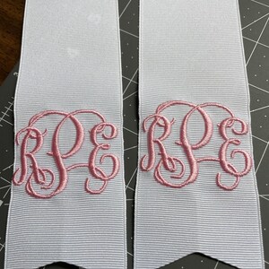 K&ouml;nnte beinhalten: Zwei wei&szlig;e Ripsbandstreifen mit einem rosa gestickten Monogramm, das die Buchstaben RPE in einer Schreibschrift zeigt. Jedes Bandende ist V-f&ouml;rmig zugeschnitten, und die B&auml;nder liegen auf einer schwarzen Schneidematte mit wei&szlig;en Gitterlinien.