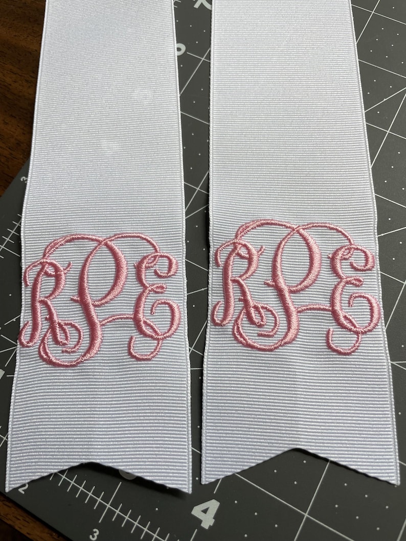 K&ouml;nnte beinhalten: Zwei wei&szlig;e Ripsbandstreifen mit einem rosa gestickten Monogramm, das die Buchstaben RPE in einer Schreibschrift zeigt. Jedes Bandende ist V-f&ouml;rmig zugeschnitten, und die B&auml;nder liegen auf einer schwarzen Schneidematte mit wei&szlig;en Gitterlinien.