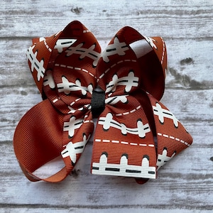 May include: A brown and white hair bow with a football pattern. The bow is tied with a black elastic band.