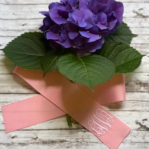 K&ouml;nnte beinhalten: Eine leuchtend violette Hortensienbl&uuml;te mit dunkelgr&uuml;nen Bl&auml;ttern, die auf einem zartrosa Band liegt. Das Band ist an einem Ende mit einem eleganten, wei&szlig; gestickten Monogramm aus drei ineinander verschlungenen Buchstaben versehen.