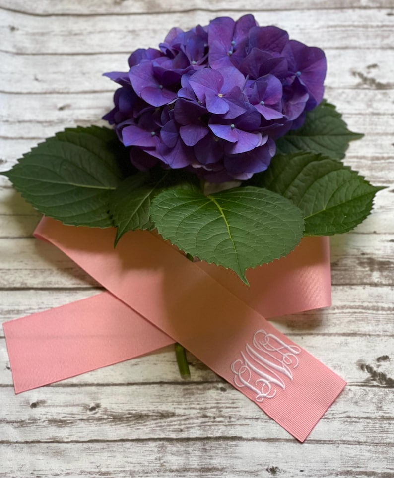 K&ouml;nnte beinhalten: Eine leuchtend violette Hortensienbl&uuml;te mit dunkelgr&uuml;nen Bl&auml;ttern, die auf einem zartrosa Band liegt. Das Band ist an einem Ende mit einem eleganten, wei&szlig; gestickten Monogramm aus drei ineinander verschlungenen Buchstaben versehen.
