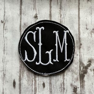 May include: Black circular patch with white embroidered initials "SLM". The patch has a white stitched border. The background is a light wood grain pattern.