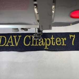 May include: A dark blue fabric strip secured in an embroidery hoop, featuring the gold embroidered text "DAV Chapter 7". The strip is positioned directly beneath the needle and presser foot of a sewing machine, which has a glowing red light.