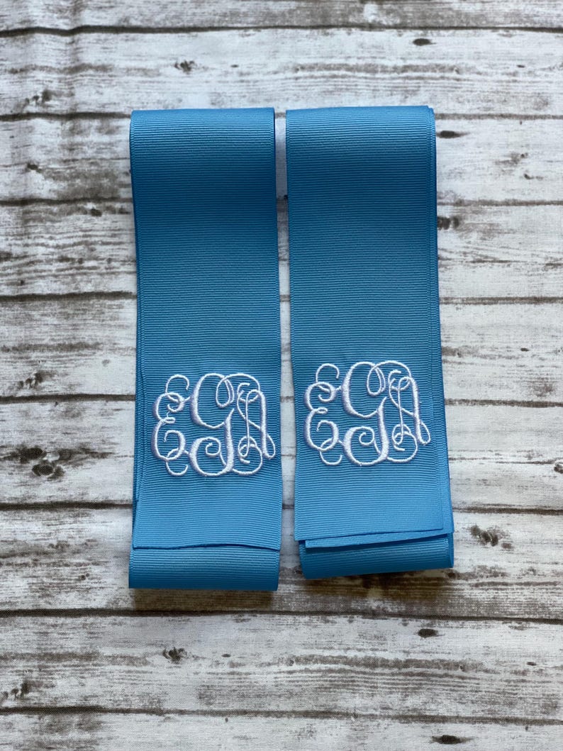 K&ouml;nnte beinhalten: Zwei Streifen aus hellblauem Ripsband, jeweils mit einem wei&szlig;en gestickten Monogramm mit den Buchstaben E, G und A in einer dekorativen Schreibschrift. Die B&auml;nder haben eine gerippte Textur und liegen nebeneinander.