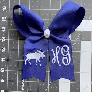 May include: A royal blue hair bow made of grosgrain ribbon with a white center knot. The left tail features an embroidered white moose silhouette, while the right tail displays white embroidered script initials. It measures 7 inches wide.