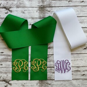 K&ouml;nnte beinhalten: Eine gr&uuml;ne Ripsbandschleife mit gold gestickten Monogramm-Initialen SPE auf den beiden herabh&auml;ngenden Enden, platziert neben einem einzelnen wei&szlig;en Ripsbandende mit einem violett gestickten Monogramm der Buchstaben AWC.