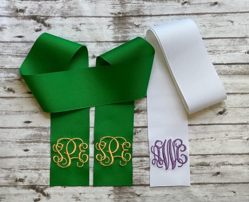 K&ouml;nnte beinhalten: Eine gr&uuml;ne Ripsbandschleife mit gold gestickten Monogramm-Initialen SPE auf den beiden herabh&auml;ngenden Enden, platziert neben einem einzelnen wei&szlig;en Ripsbandende mit einem violett gestickten Monogramm der Buchstaben AWC.