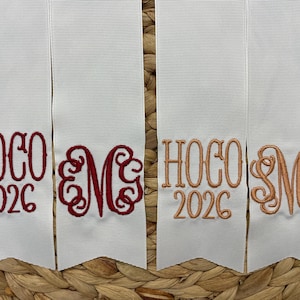 May include: Four white ribbons with pointed ends, each featuring embroidery. Two ribbons display the text HOCO 2026 in red and copper thread. The other two ribbons feature ornate, swirling monogram letters in matching red and copper embroidery.