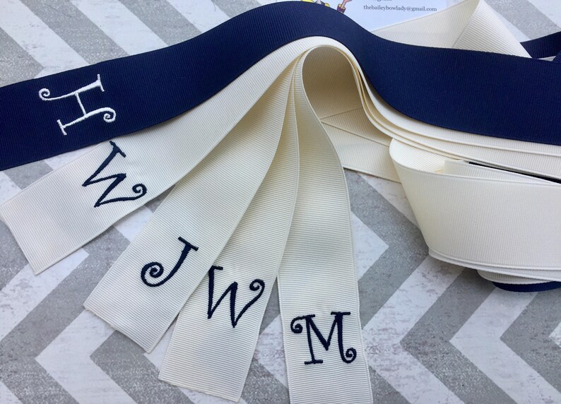 May include: Five white satin ribbons with navy blue embroidered letters, H, W, J, W, and M. The ribbons are arranged in a fan shape with the navy blue ribbon on top.