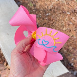 May include: A large, bright pink hair bow with a yellow center. The bow features an embroidered design of a yellow sun, a blue heart, and blue waves. The bow is held in a hand.