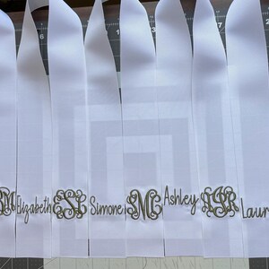 K&ouml;nnte beinhalten: Eine Reihe von wei&szlig;en Satinb&auml;ndern, die nebeneinander angeordnet sind. Jedes Band ist mit einem personalisierten Namen oder einem dekorativen dreibuchstabigen Monogramm in dunkelgoldfarbenem Metallic-Garn bestickt. Die B&auml;nder liegen flach auf einer dunklen Schneidematte.