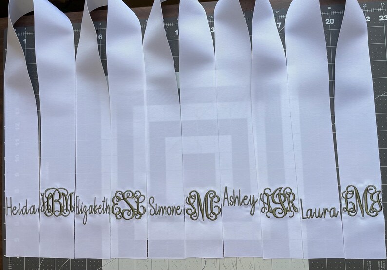 K&ouml;nnte beinhalten: Eine Reihe von wei&szlig;en Satinb&auml;ndern, die nebeneinander angeordnet sind. Jedes Band ist mit einem personalisierten Namen oder einem dekorativen dreibuchstabigen Monogramm in dunkelgoldfarbenem Metallic-Garn bestickt. Die B&auml;nder liegen flach auf einer dunklen Schneidematte.