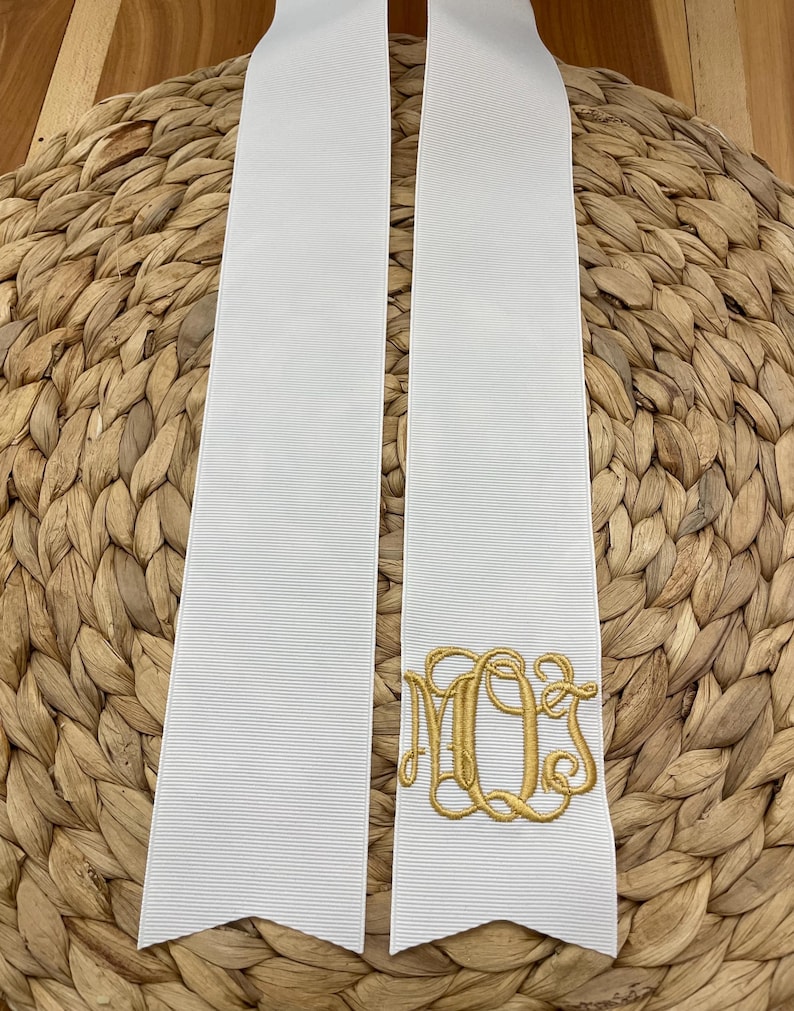 K&ouml;nnte beinhalten: Zwei lange, wei&szlig;e, gerippte Stoffb&auml;nder mit spitz zulaufenden Enden. Auf einem der B&auml;nder befindet sich ein goldfarbenes, gesticktes Monogramm mit den ineinander verschlungenen Buchstaben M, Q und S in dekorativer Schreibschrift am unteren Rand.