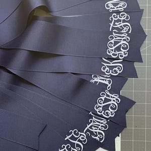 K&ouml;nnte beinhalten: Eine Sammlung marineblauer B&auml;nder mit wei&szlig; gestickten Monogrammen in Schreibschrift. Die B&auml;nder sind f&auml;cherf&ouml;rmig auf einer Schneidematte mit sichtbaren Zentimeter-Markierungen angeordnet und zeigen die detaillierte Stickerei der Buchstaben.