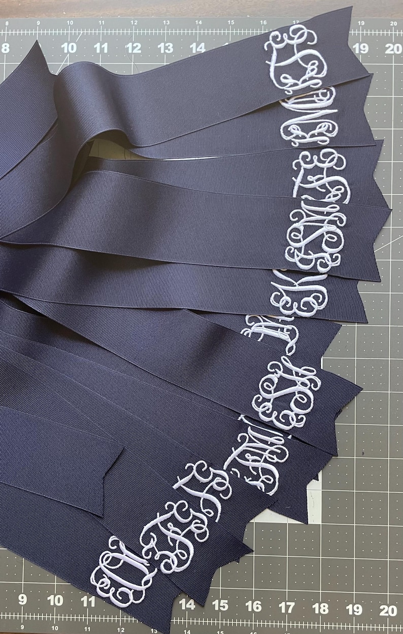 K&ouml;nnte beinhalten: Eine Sammlung marineblauer B&auml;nder mit wei&szlig; gestickten Monogrammen in Schreibschrift. Die B&auml;nder sind f&auml;cherf&ouml;rmig auf einer Schneidematte mit sichtbaren Zentimeter-Markierungen angeordnet und zeigen die detaillierte Stickerei der Buchstaben.