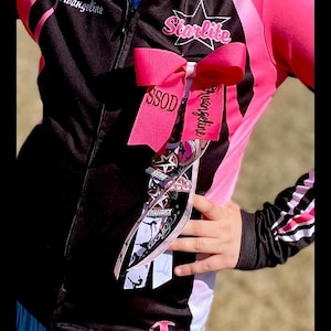 May include: A black and pink zip-up jacket with the text Starlite, Avangeline, and SSOD. A large pink hair bow with long ribbons hangs from the jacket, featuring various star-themed graphics, the name Avangeline, and silhouettes of dancers.