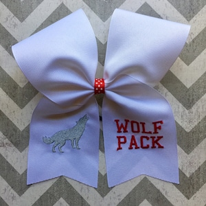 May include: A white cheer bow with a red and white polka dot ribbon in the centre. The left side of the bow has a grey embroidered wolf howling. The right side of the bow has the words "WOLF PACK" embroidered in red.