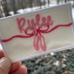 May include: Embroidered patch with the name "Rylee" in pink script, above a pink ribbon and ballet shoes. The patch is rectangular, with a cream-colored background and white border. The design is stitched onto the patch.