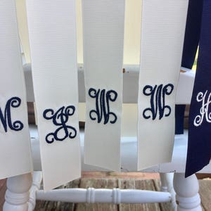 May include: Five white satin ribbons with navy blue embroidered monograms. The monograms are "M", "S", "W", "W", and "H".