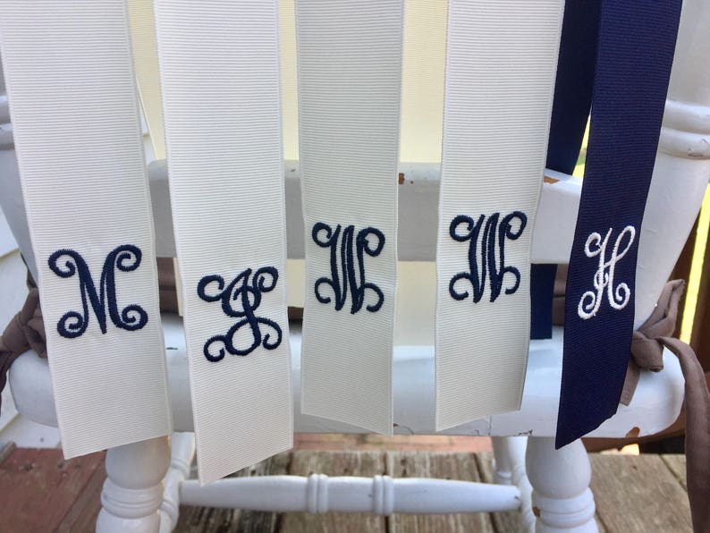 May include: Five white satin ribbons with navy blue embroidered monograms. The monograms are "M", "S", "W", "W", and "H".