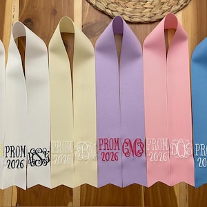 May include: A collection of graduation sashes in various colors, including white, cream, lavender, pink, and blue. Each sash is embroidered with "PROM 2026" and a monogram. The sashes are displayed against a wooden background.