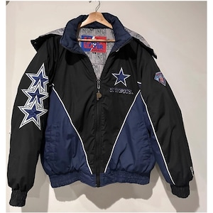 May include: A black and navy blue Dallas Cowboys NFL jacket with a full-length zipper, a high collar, and a detachable hood. The jacket features three large blue and white star patches on the right sleeve, a star logo on the chest, and NFL branding.