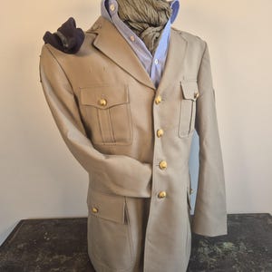 Op de afbeelding: Een beige jas in militaire stijl met goudkleurige knopen, vier voorzakken en epauletten. De jas wordt gedragen over een lichtblauw overhemd met kraag en een gestructureerde olijfgroene sjaal, met een donkerblauwe baret op de schouder.