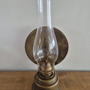 Op de afbeelding: Een koperen olielamp met een heldere glazen cilinder en een ronde reflector aan de achterzijde. De voet van de lamp is voorzien van het woord HORNET in reliëf. De metalen onderdelen hebben een verweerde afwerking en een lont is zichtbaar.