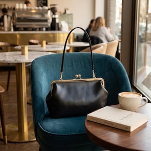 Könnte beinhalten: Eine schwarze Handtasche aus Leder mit einem goldfarbenen Metallrahmen und einem Schnappverschluss. Die Tasche verfügt über einen einzelnen dünnen schwarzen Henkel, der mit goldener Hardware am Rahmen befestigt ist. Das Design ist strukturiert.