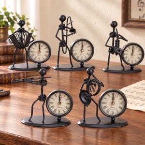May include: A set of five black metal desk clocks, each featuring a stylized musician figure playing an instrument. The round clock faces have cream-colored backgrounds with black Roman numerals and quartz movement, mounted on oval black metal bases.