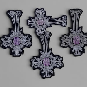 Könnte beinhalten: Vier gestickte Aufnäher mit einem silbernen und violetten Kreuzdesign auf schwarzem Hintergrund. Jeder Aufnäher hat ein komplexes, symmetrisches Muster mit einem zentralen violetten Quadrat und verlängerten Armen mit Schnörkeln.