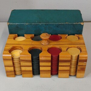 May include: A wooden poker chip rack with a central handle containing stacks of yellow, black, and red circular chips. Behind the rack sits a rectangular teal storage box with a worn finish. The rack features eight vertical slots for chip storage.