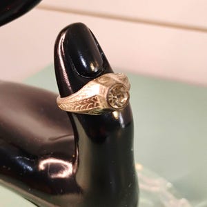 Pode incluir: Um anel de metal prateado com uma pedra redonda transparente facetada engastada numa moldura circular. A banda apresenta um padrão gravado intrincado de folhas ou penas nas laterais, estendendo-se da cravação central para a parte de trás.