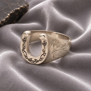 Puede incluir: Un anillo de metal plateado con forma de herradura. La parte frontal de la herradura presenta un patrón de enredadera grabado con hojas pequeñas, mientras que los lados de la banda del anillo muestran un diseño texturizado de una cabeza de caballo.