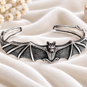 May include: A silver-toned metal cuff bracelet featuring a central bat motif with outstretched wings. The design includes detailed textures on the bat's body and wings, with an open-ended band structure that allows for adjustment around the wrist.