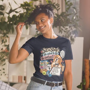May include: A dark navy blue t-shirt featuring a graphic of a golden retriever wearing glasses and a bow tie, holding a pen over a contract. The text reads "Sit & Stay? He asked to see the contract!" with illustrations of books, a mug, and dog toys.