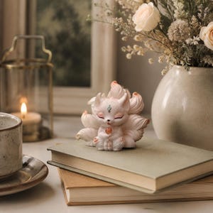 May include: A small, white, multi-tailed fox figurine with soft pink accents on the ears and tail tips. The fox has closed eyes, a small teal forehead marking, and holds a smooth, translucent orange sphere between its front paws.