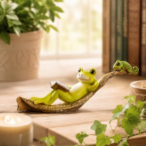 May include: A lime green frog figurine reclining on a textured brown leaf-shaped base whilst holding a small brown book. A tiny green chameleon is perched on the tip of the leaf. The frog has large black eyes and a smooth, light-coloured belly.