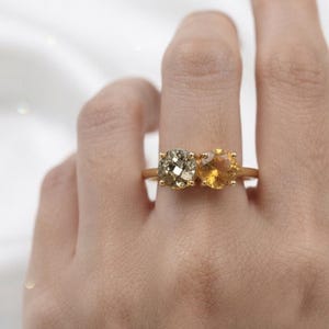 Pode incluir: Um anel de ouro com duas pedras de corte redondo lado a lado. Uma pedra é uma pirita metálica e reflexiva com superfície facetada, enquanto a outra é um citrino amarelo brilhante e translúcido. Ambas as pedras estão fixas numa banda de ouro.