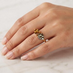 May include: A gold ring featuring two pear-shaped gemstones set side-by-side. One stone is a translucent amber-coloured citrine, and the other is a dark, metallic-toned pyrite. The stones are held in place by a thin, polished gold metal band.
