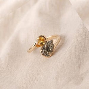 May include: A gold bypass ring featuring two pear-shaped settings. One setting contains a faceted yellow gemstone, while the other holds a dark, textured druzy stone. The band is smooth and polished, creating a continuous, flowing design.