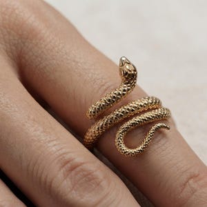 Puede incluir: Un anillo dorado con forma de serpiente enroscada, que presenta una superficie texturizada que imita las escamas. La cabeza de la serpiente está situada en la parte superior, mientras que su cuerpo se enrolla alrededor del dedo en espiral.