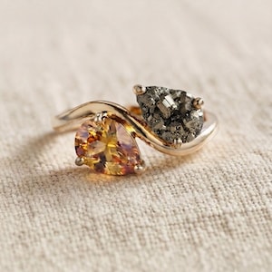 May include: A bypass ring crafted from a warm-toned metal, featuring two pear-shaped stones. One stone is a faceted, translucent amber-coloured gem, while the other is a textured, metallic grey pyrite stone with a crystalline surface structure.