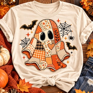 Puede incluir: Una camiseta de manga corta color crema con un gráfico central de un fantasma hecho de varios patrones de patchwork en naranja, blanco y marrón. El diseño incluye murciélagos negros, telarañas, una pequeña araña y estrellas naranjas.