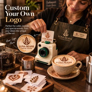 Puede incluir: Una impresora de café blanca con un anillo de lente verde, utilizada para crear arte latte. Cuenta con una ranura para plantillas cuadradas que muestran el logotipo de Luna Beans Coffee House. Una taza de cerámica con el logo en la espuma descansa sobre un plato.