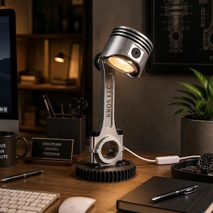 Peut inclure: Une lampe de bureau fabriquée à partir d'un piston et d'une bielle de moteur en métal argenté, montée sur une base en engrenage circulaire noir. La bielle porte l'inscription BROS LLC gravée verticalement. La lampe émet une lumière chaude.